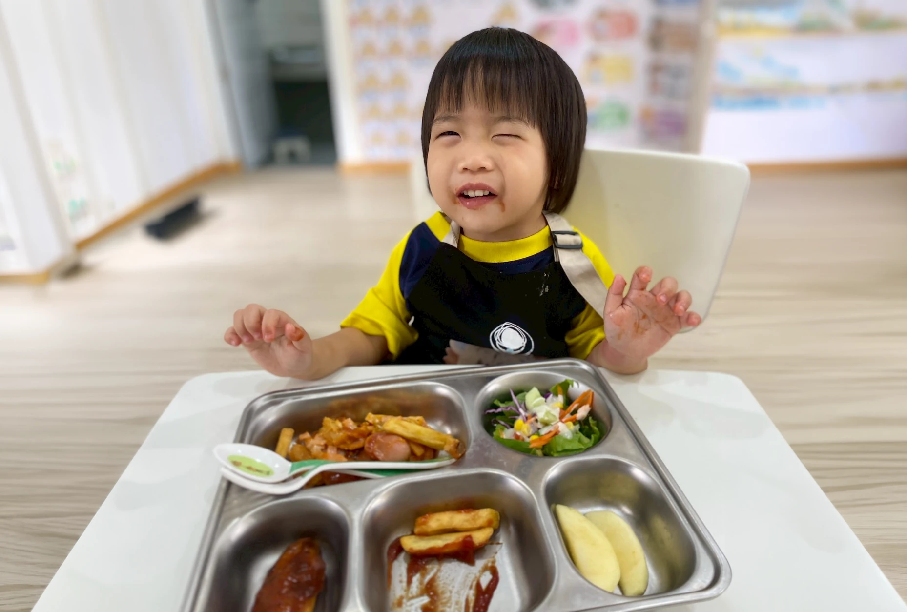 A child smiling behind a full meal tray of rice, salad, fries and fruit