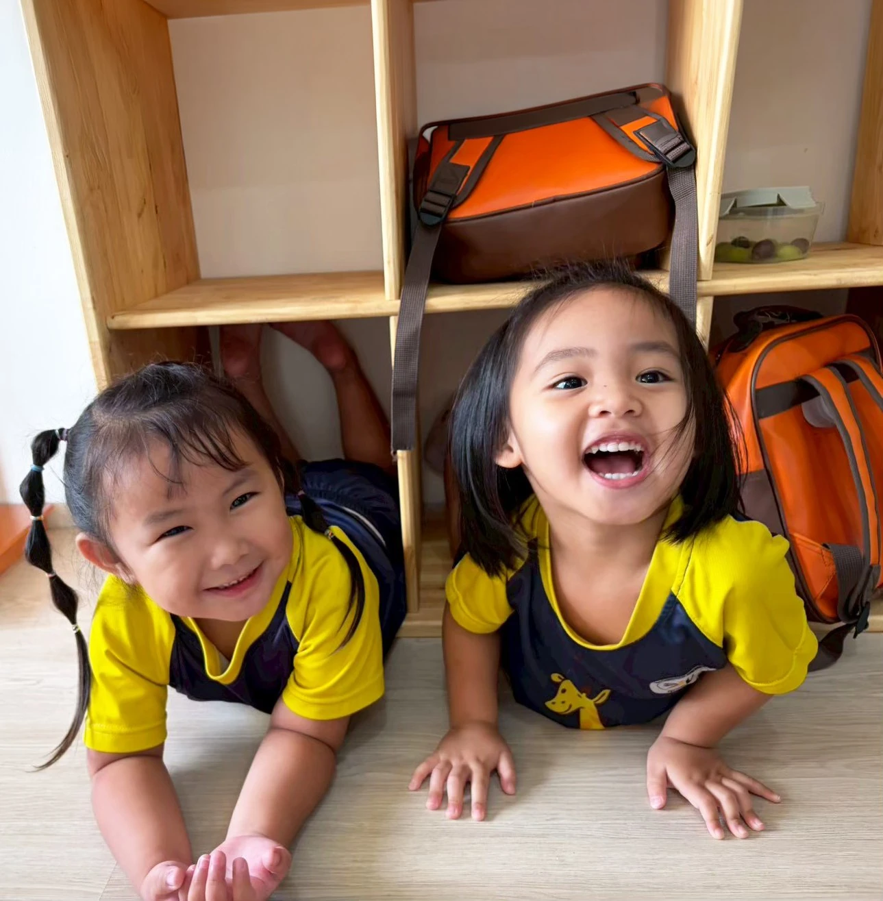 两个 Kindergarten 孩子躺在储物架下方的教室地板上，抬头对着镜头大笑