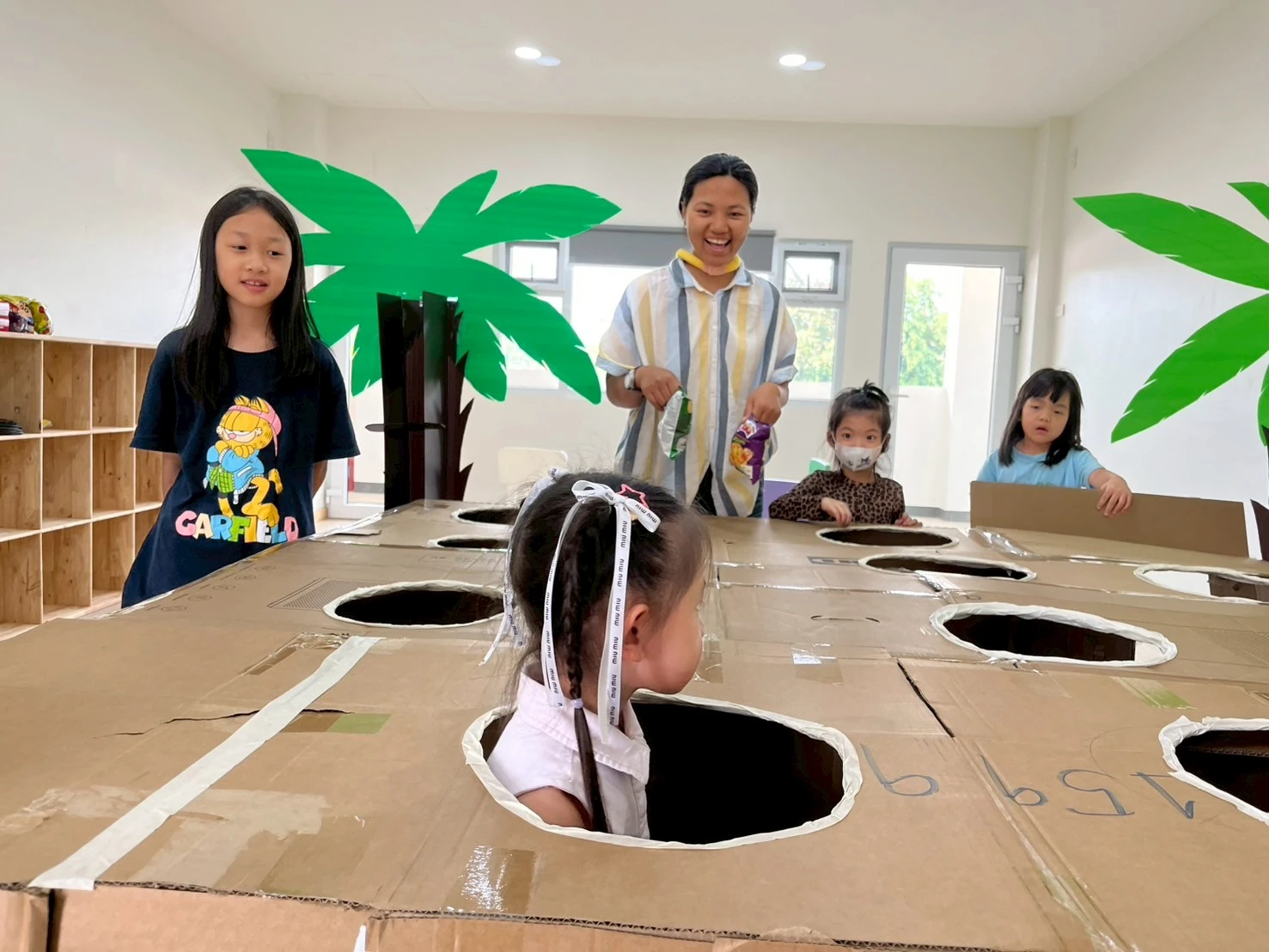 A teacher and three children gathered around a cardboard playhouse with cut-out windows, one child peeking through