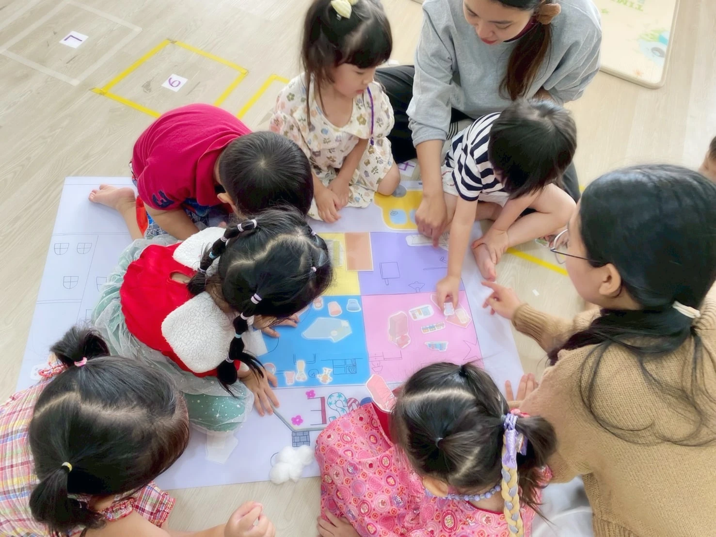 A group of children and a teacher gathered around a shared floor-map project, pointing and working together