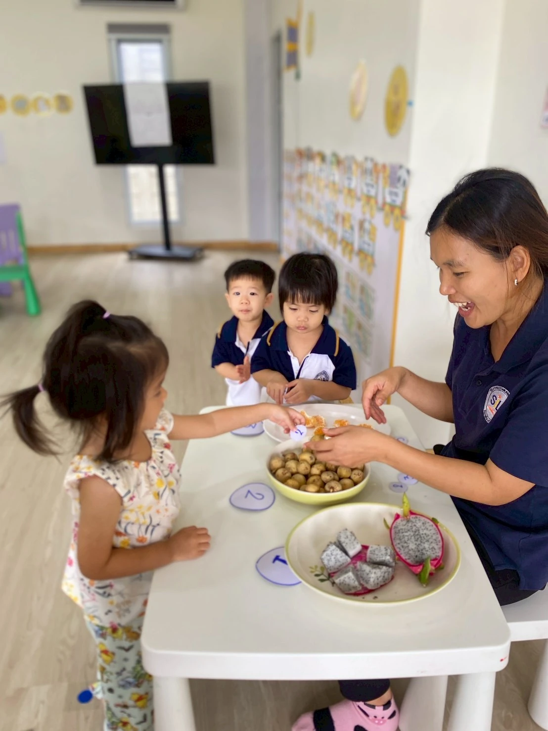 Nursery 孩子们围在装有龙眼和火龙果的碗前，老师微笑着
