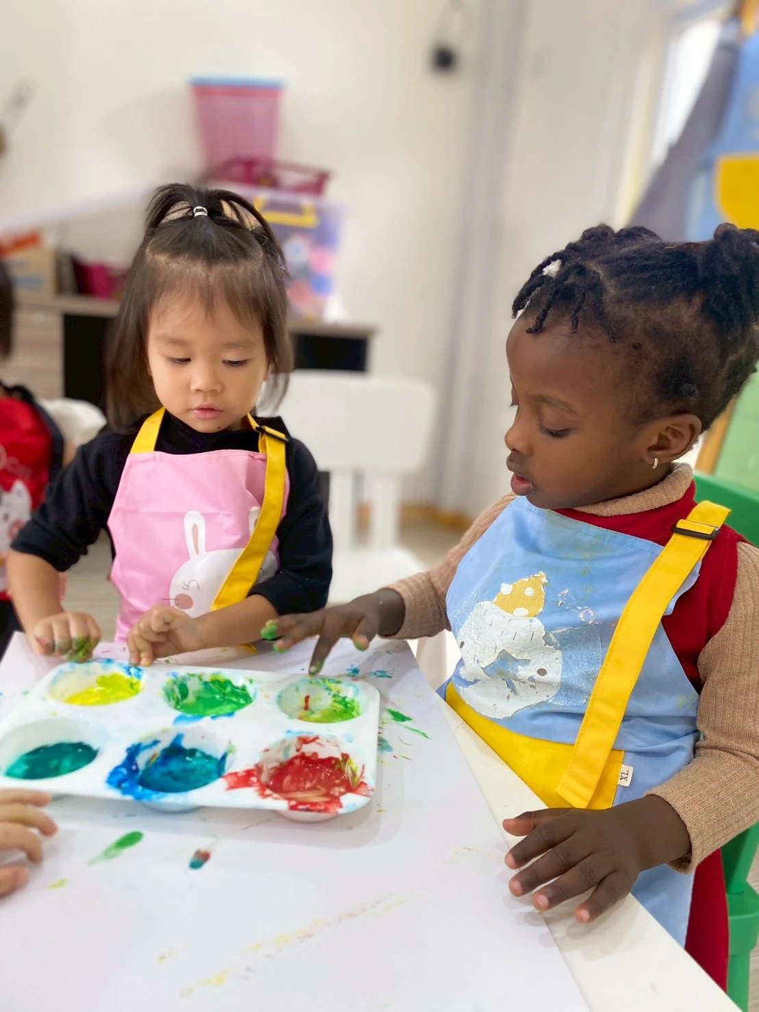 两个穿着围裙的 Nursery 孩子在桌前一起玩手指画