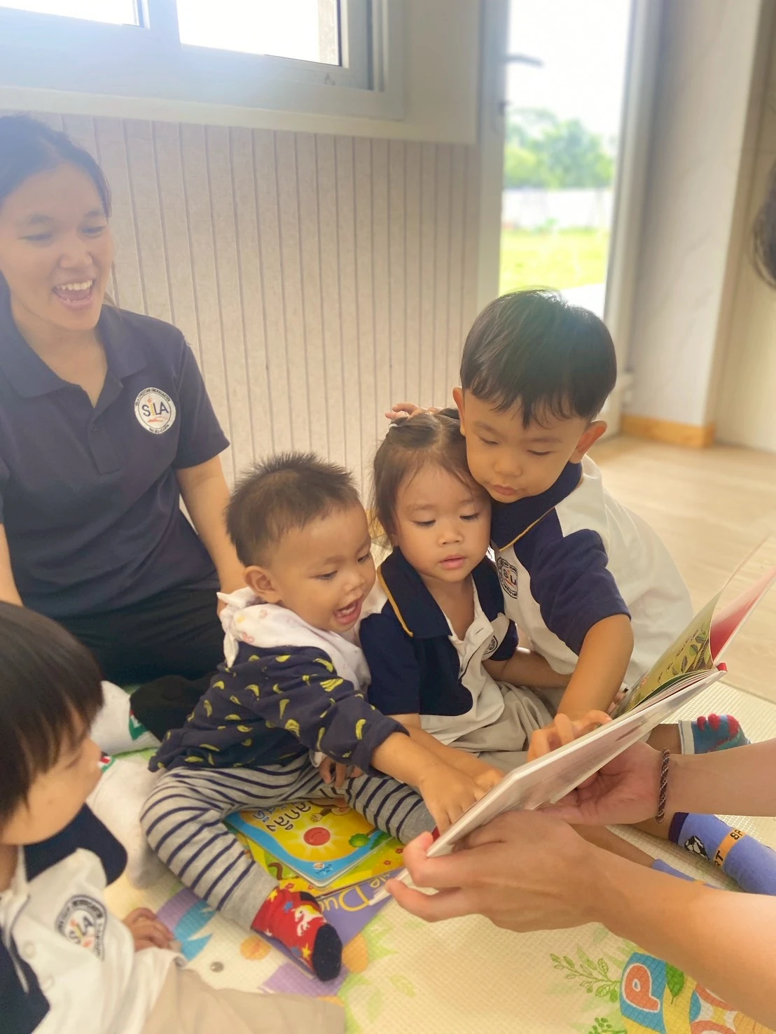 三个 Nursery 孩子和老师一起靠近看一本图画书